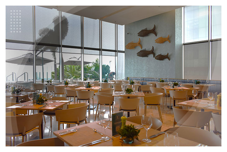 Restaurante con mesas de madera, sillas claras, ventanales altos que permiten la entrada de luz y detalles decorativos inspirados en el mar, ubicado en Estelar Cartagena de Indias Hotel