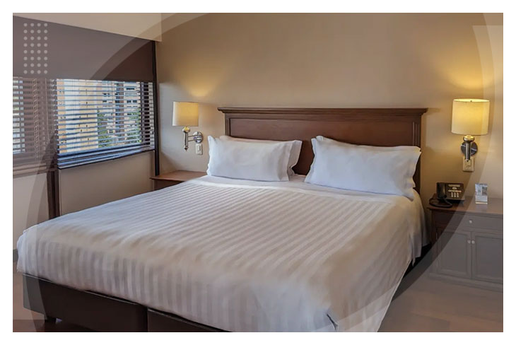 Habitación con cama amplia y sábanas blancas, ambientada con luz cálida, un par de mesas de noche, lámparas modernas y ventana con vista a la ciudad, ideal para quienes buscan promociones para reservar en el Hotel La Fontana