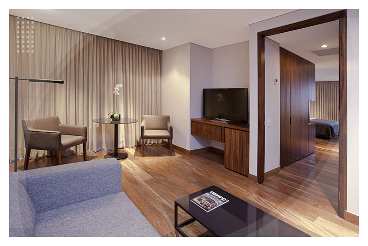 Sala de habitación con piso en madera, sofá gris, mesa de centro, televisor y un rincón acogedor con sillas y cortinas largas es parte de los espacios modernos del Hotel Estelar del Parque de la 93