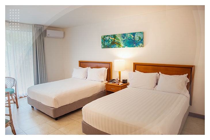 Habitación con dos camas, decoración sencilla en tonos claros, mesa de noche con lámpara y acceso a un balcón, en un hotel cerca a la playa Pozos Colorados