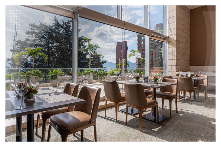 Restaurante iluminado con múltiples sillas y mesas junto a grandes ventanales que ofrecen una vista panorámica de Medellín, ideal para disfrutar del hospedaje en el Poblado de Medellín con descuento