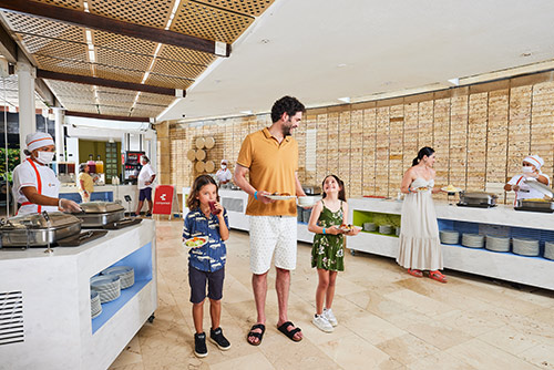 Un hombre, una mujer, un niño y una niña, que visten ropa de clima cálido con colores azul oscuro, blanco, verde musgo y amarillo mostaza, están de pie sosteniendo platos en el área  de comidas de Lagomar, a su alrededor hay mostradores, bandejas y varios cocineros