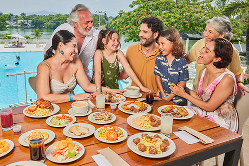 Dos hombres, tres mujeres y dos niños, que visten ropa de clima cálido de colores blanco, beige, verde musgo, mostaza, azul oscuro, amarillo y rosado, están sentados frente a una mesa de madera en la cual hay 16 platos de comida y 5 bebidas ofrecidas en el Hotel Lagomar
