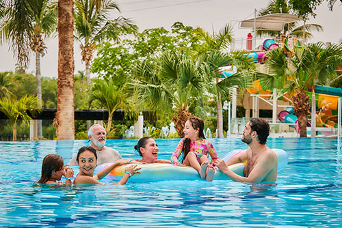 Dos hombres, dos mujeres y dos niñas, que visten trajes de baño, están dentro de una piscina en Lagomar. Una de las niñas está sentada sobre un flotador de cama de color blanco y naranja, mientras las demás personas se divierten a su alrededor y disfrutan de los hoteles Compensar
