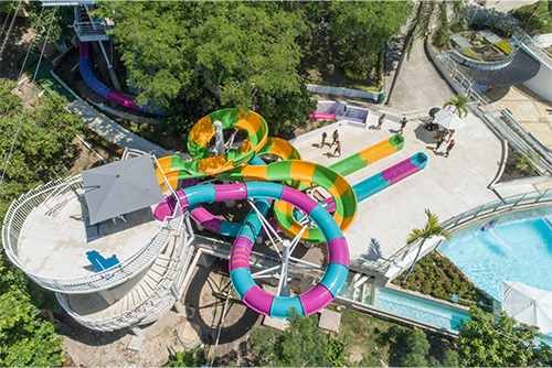 Plano cenital de dos toboganes en espiral, el primero de color azul y morado y el segundo amarillo y verde, junto a ellos hay ocho personas caminando alrededor del hotel y parque Lagosol