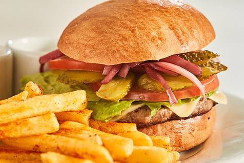 Hamburguesa con carne, queso, lechuga, tomate, jamón y pepinillos, junto a papas fritas sobre un plato blanco