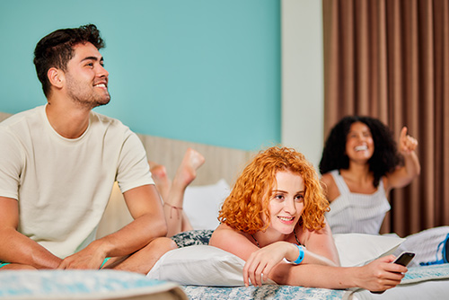 Una mujer de cabello cobrizo, que viste ropa de color azul oscuro, está acostada boca abajo en una cama del Hotel Lagomar, sosteniendo un control negro en su mano, a su lado derecho hay un hombre sentado, que viste de color beige y cian, y a su lado izquierdo hay una mujer sentada señalando con su mano hacia el frente