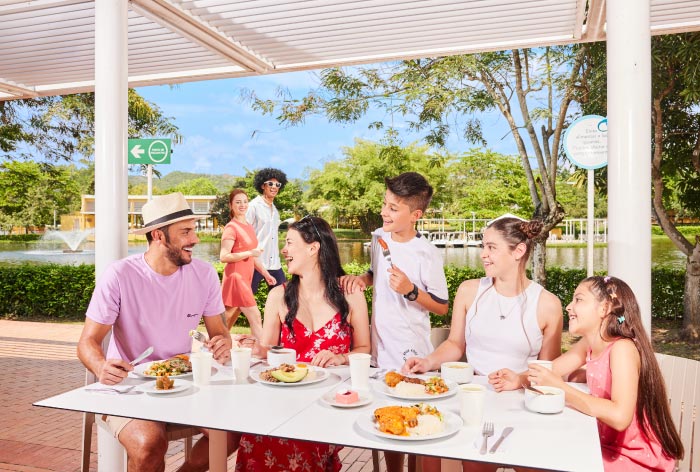 Familia sentada en un comedor de madera de color blanco, comparten varios alimentos y sonríen, mientras disfrutan de las mejores tarifas y planes del hotel Lagosol Compensar. En el fondo se logra ver un lago, gran cantidad de vegetación y el cielo azul