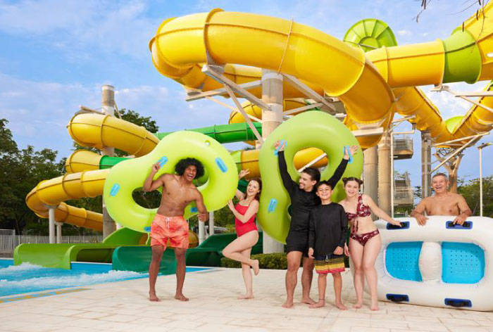 Seis personas de diferentes edades están frente a un gran tobogán de color verde y amarillo, celebrando y sonriendo, durante una de las actividades incluidas con los planes vacacionales en Lagosol. En el fondo se logra ver vegetación y el cielo con algunas nubes