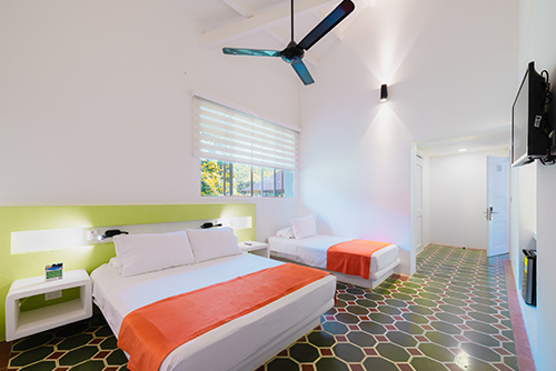 Habitaciones en el centro vacacional Las Palmeras con baldosas oscuras y geométricas y dos camas, una doble y otra personal de tendidos blancos y naranjas, acompañadas de un ventilador, un televisor y dos mesas de noche