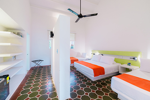 Habitación con tarifas especiales del centro vacacional Las Palmeras que tiene un piso de baldosas verde oliva con púrpura, tres camas dobles con tendidos blancos y naranjas, cada una al lado de una mesa de noche