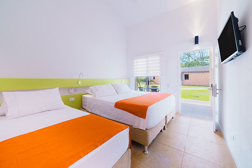 Habitación del plan básico de acomodación doble para adultos que cuenta con dos camas dobles de sábanas blancas y naranjas, un televisor y una ventana que hace parte de las tarifas especiales del centro vacacional Las Palmeras