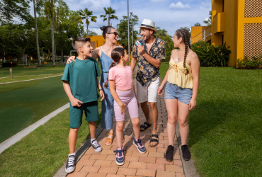Familia de cinco personas que usan ropa cómoda están caminando por un sendero que está rodeado de edificios con paredes de color mostaza en donde quedan las habitaciones del hotel Lagosol, las cuáles están rodeadas de naturaleza