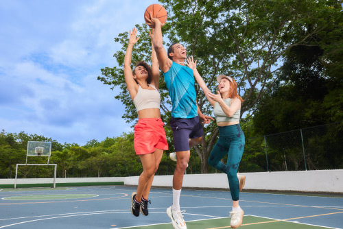 Dos mujeres y un hombre, que usan ropa deportiva, están en una cancha de baloncesto saltando tratando de encestar el balón en el aro de uno de los escenarios deportivos de los planes vacacionales en Lagosol