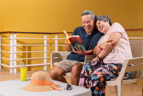 Pareja de adultos mayores sentados en una silla de color beige, en un salón de paredes de color mostaza, está leyendo un libro en una de las locaciones incluidas con el hospedaje en el hotel Lagosol