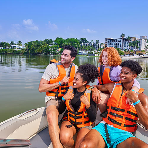 Grupo de amigos disfrutando de uno de los planes turísticos del hotel Lagomar fin de semana festivo, en el cual están sentados en un bote, dos hombres y dos mujeres en pareja, cada uno con chaleco salvavidas, mientras sonríen y disfrutan la vista del lago. En el fondo se pueden ver las instalaciones del hotel y gran cantidad de árboles alrededor