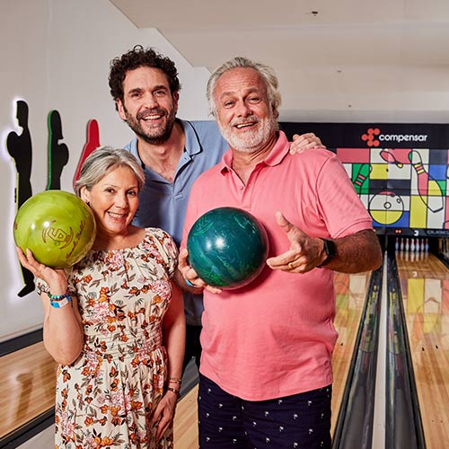 Tres personas jugando bolos aprovechando el plan fin de semana festivo en hotel Lagomar. Dos son  adultos mayores, un hombre vestido de camiseta polo rosada y pantalón azul oscuro con puntos blancos, la mujer con vestido de flores