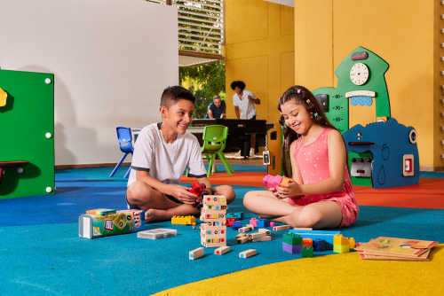 Dos niños, sentados en un piso de diferentes colores están jugando Jenga, en un salón que tiene paredes de color blanco y mostaza a las que se puede acceder luego de adquirir uno de los planes vacacionales en Lagosol