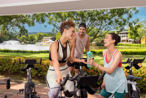 Tres personas jóvenes están en un gimnasio que tiene bicicletas de spinning, en un lugar rodeado de naturaleza y de un lago, al cual que se puede acceder luego de hacer una reserva en Lagosol Compensar