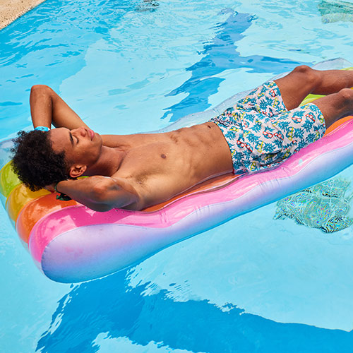 Un hombre, que usa una pantaloneta de color blanco con estampado azul, amarillo y rojo, está acostado sobre un flotador con forma de cama de colores azul, rosado, naranja, amarillo y verde, relajándose en una piscina del Hotel Lagomar