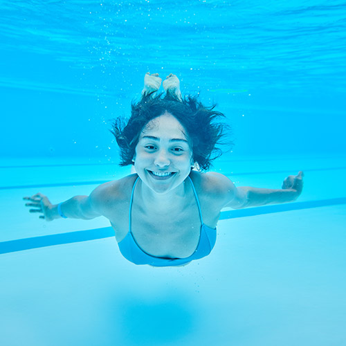Una mujer de cabello corto, que usa un vestido de baño de color azul, sonríe y extiende los brazos hacia atrás mientras está sumergida en una piscina del Hotel Lagomar