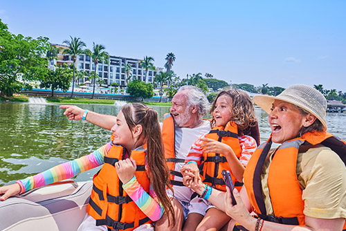 Una mujer mayor, un hombre mayor, un niño y una niña, que usan chalecos salvavidas de color naranja y negro, ropa de clima cálido en tonos blanco, amarillo, rojo, rosado y azul, están sentados sobre un bote de color gris, señalando y divirtiéndose con uno de los planes turísticos  en Lagomar, a su alrededor hay gran variedad de vegetación