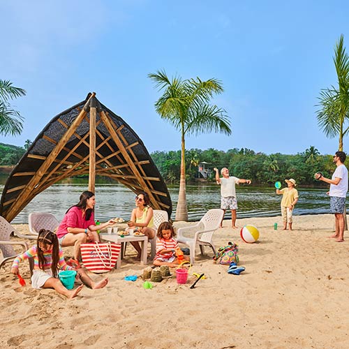 Dos mujeres, que visten de colores fucsia y amarillo, están sentadas bajo un parasol de madera en la playa disfrutando de una de los planes turísticos en Lagomar, frente a ellas hay un niño y una niña jugando sentados en la arena. Detrás de ellos hay una mujer y dos hombres de pie, vestidos con ropa de colores amarillo, blanco y azul