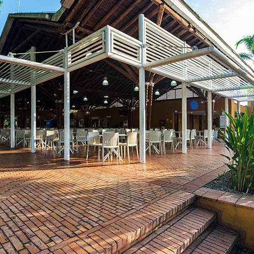 Gran estructura metálica que cubre varias sillas y mesas, debajo de la que se puede ver el piso de ladrillo. En el exterior hay vegetación que se puede conocer al acceder a las promociones en Lagosol temporada alta