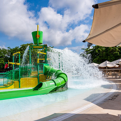 Gran tobogán verde con amarillo, ubicado en la mitad de una piscina, está salpicando agua hacia los lados, en donde hay varios parasoles blancos que cubren mesas y sillas en donde se puede disfrutar de la vista y de los mejores planes exclusivos en el hotel Lagosol. Al fondo se ven algunos árboles que rodean el lugar