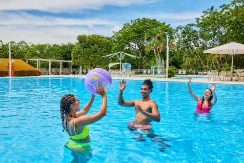 Tres personas jóvenes, que usan vestido de baño, están jugando dentro de una piscina de agua cristalina con un balón luego de acceder a uno de los mejores planes vacacionales en Lagosol. En el fondo se pueden ver sillas y mesas que tienen grandes parasoles blancos y gran cantidad de vegetación