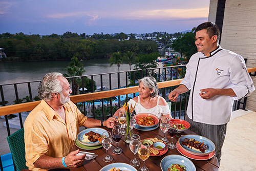 Pareja de adultos mayores disfrutando de una cena, con vista al lago, mientras son atendidos por uno de los meseros, el cual les explica los beneficios de comprar con las tarifas del plan temporada alta en hotel Lagomar