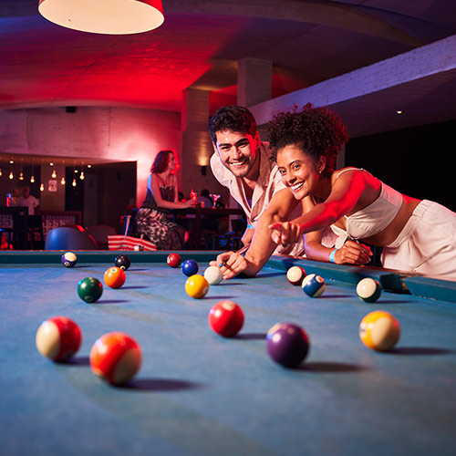 Pareja joven vestida de blanco jugando pool en un ambiente con luces de neón en el hotel en el que compraron el plan temporada alta Lagomar. Al fondo se ve a una mujer disfrutando de un coctel junto a un gran ventanal que tiene una extensión de luces cálidas