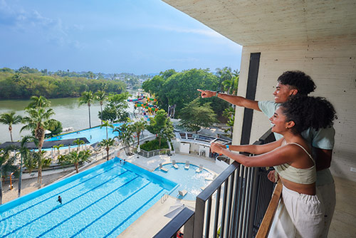 Pareja mirando una piscina y un lago desde un balcón de gran altura, sonriendo y disfrutando de la vista a la cual pueden acceder con las ofertas de temporada alta en Lagomar. En el fondo se alcanza a ver gran cantidad de vegetación y un cielo azul