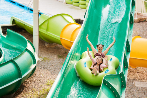 Dos niñas que están descendiendo por un tobogán de colores verde y amarillo, levantan los brazos y sonríen luego de acceder a uno de los mejores planes turísticos de Lagosol