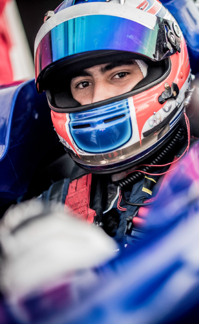 Un piloto con casco azul y traje deportivo ajusta su volante dentro del monoplaza, preparándose para la salida del pit; la imagen captura la intensidad que rodea los viajes a Barcelona para el gran premio de Cataluña, en medio del equipo técnico