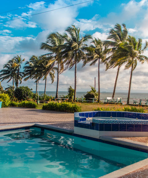 Primer plano de piscina de agua cristalina que esta al aire libre junto a una zona de jacuzzi con acabados en baldosa azul. Al fondo, palmeras y las olas del mar, que se pueden disfrutar c con los planes viajando y cuidando ando de Compensar
