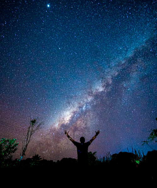 Persona con los brazos extendidos hacia el cielo, admirando un paisaje estrellado donde resalta la Vía Láctea, en medio de la naturaleza. Tonos azules, rosados y blancos se combinan en el firmamento, una experiencia única que se puede vivir al acceder a los planes de turismo ecológico en Colombia