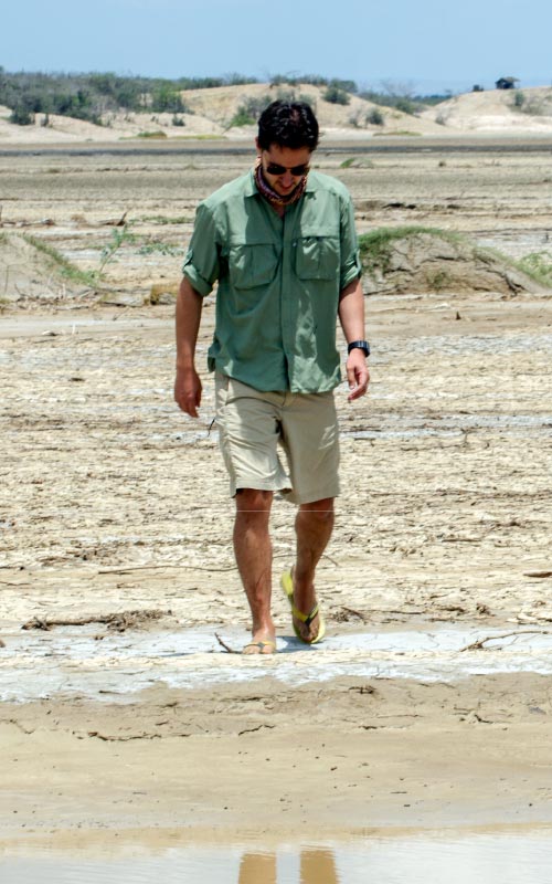 Hombre con camisa verde y pantalón claro caminando solo por una playa desértica de La Guajira, rodeado de arena y formaciones secas